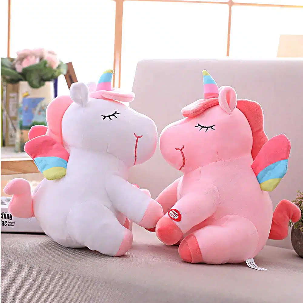PELUCHE LICORNE LUMINEUSE ROSE AILÉE 21 DEUX PELUCHES LICORNES LUMINEUSE BLANCHE ET ROSE AILÉE SUR UN CANAPÉ