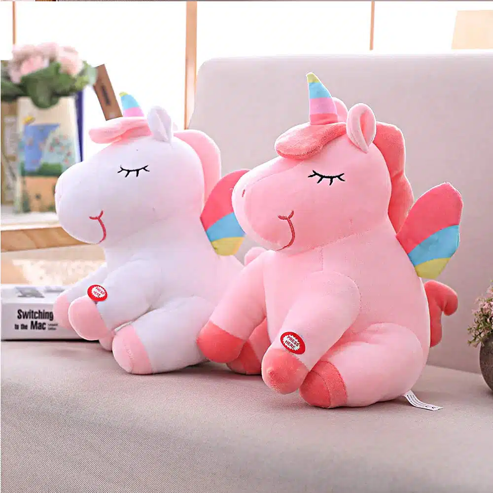 PELUCHE LICORNE LUMINEUSE ROSE AILÉE 22 DEUX PELUCHES LICORNES LUMINEUSE BLANCHE ET ROSE AILÉE SUR UN CANAPÉ VU DE COTÉ