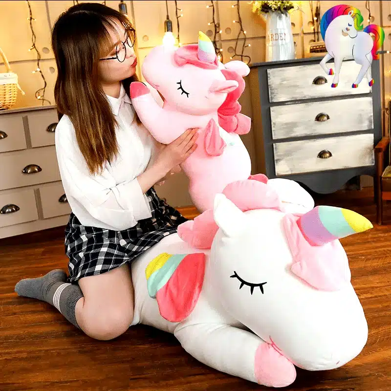 PELUCHE LICORNE GÉANTE ROSE AILÉE 16 JEUNE FILLE SUR UNE PELUCHE LICORNE GÉANTE BLANCHE AILÉE QUI TIENT UNE PELUCHE LICORNE GÉANTE ROSE AILÉE
