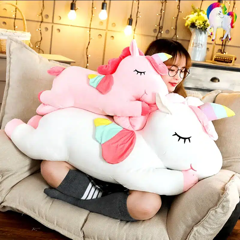 PELUCHE LICORNE GÉANTE ROSE AILÉE 7 JEUNE FILLE SUR UN FAUTEUIL AVEC UNE PELUCHE LICORNE GÉANTE BLANCHE AILÉE ET UNE PELUCHE LICORNE GÉANTE ROSE AILÉE